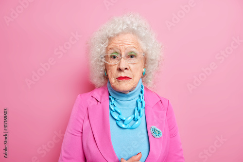 Portrait of serious mature grey haired woman dressed in elegant clothes with jewelry wears makeup remains beautiful despite her old age poses indoor against pink background. Fashionable grandmother