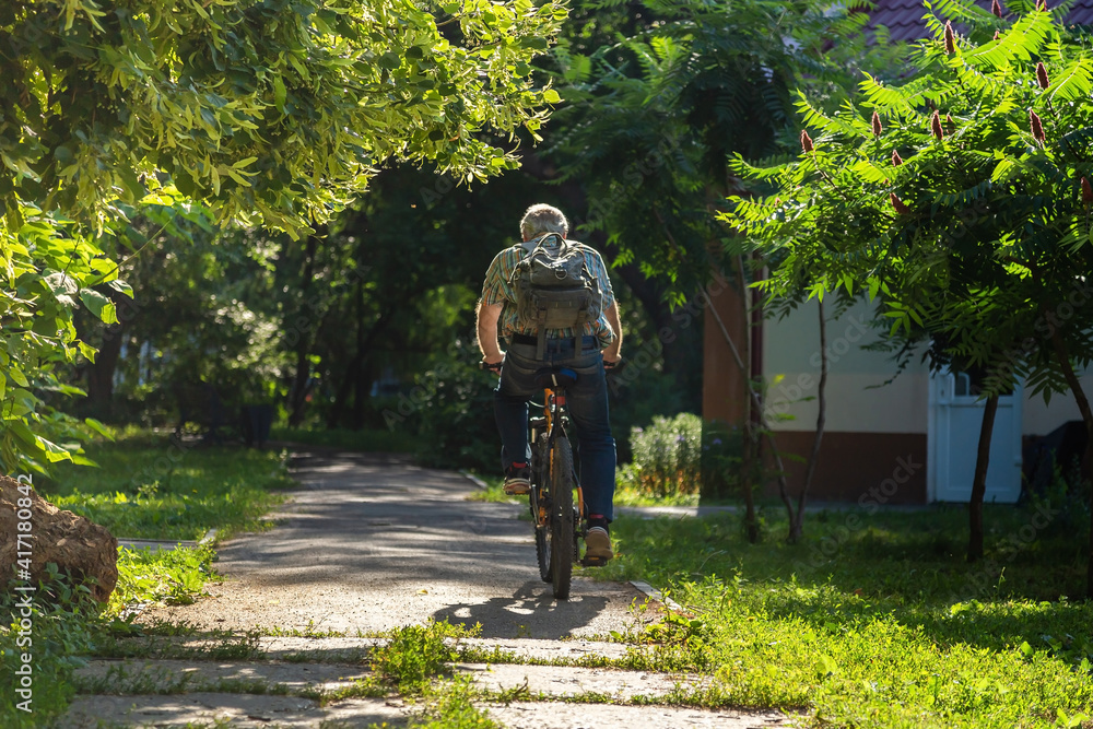 Fototapeta premium Elderly man european leads an active healthy lifestyle while riding a bicycle in the park in summer, view from the back