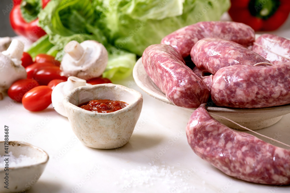 Raw uncooked italian sausages salsiccia in plate on white marble table. Green salad, vegetables and tomato sauce around.
