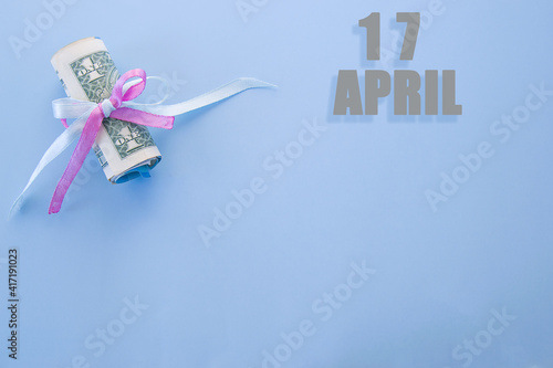calendar date on blue background with rolled up dollar bills pinned by blue and pink ribbon with copy space. April 17 is the seventeenth day of the month