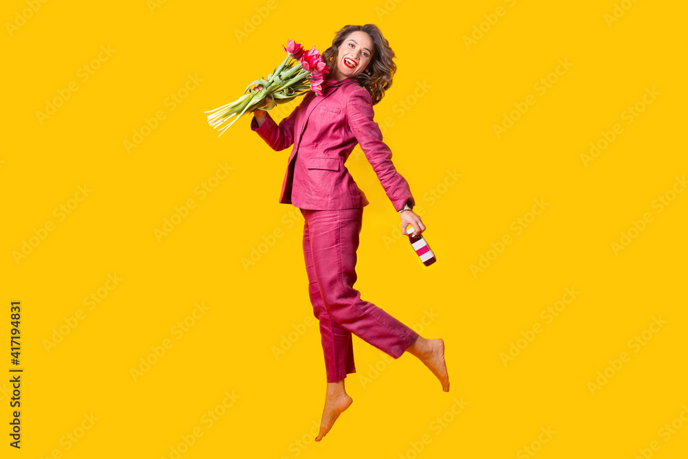 Photo of happy woman in suit jumping over yellow background and holding bunch of tulips and fresh bottle juice.