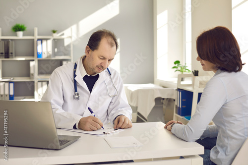 Healthcare in a hospital or private clinic. Middle-aged doctor writes a prescription to a female patient in his hospital office. Concept of disease prevention and consultations in the medical clinic.