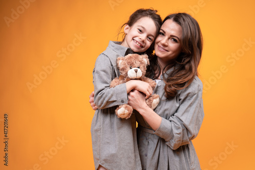 Mom holding a toy bear and daughter in pigtails hug and look at the camera. Family holiday