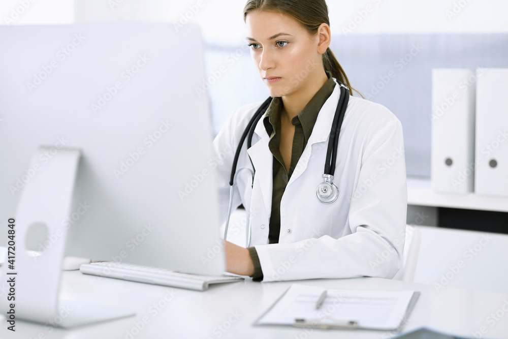 Woman-doctor typing on pc computer while sitting at the desk in hospital office. Physician at work. Data in medicine and healthcare Woman-doctor typing on pc computer while sitting at the desk in hospital office. Physician at work. Data in medicine and healthcare