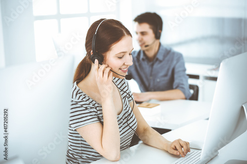 Casual dressed young woman using headset and computer while talking with customers online. Group of operators at work. Call center, business concept