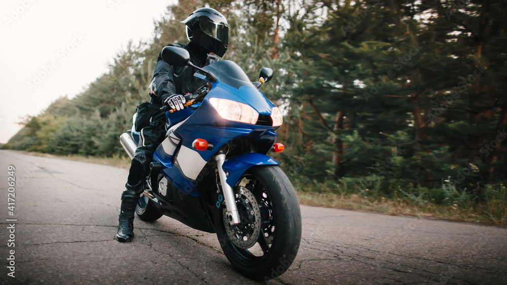 Motorcyclist in leather protective suit and black helmet sits on sports motorcycle. Biker in black rides on the road against the background of the forest