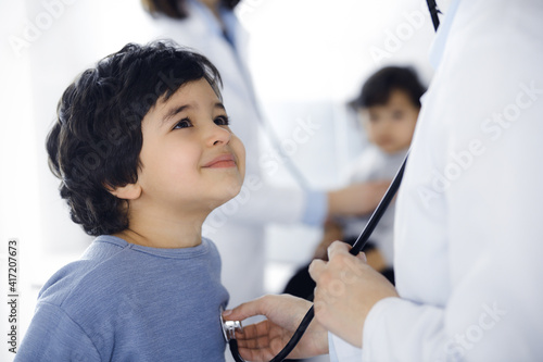 Doctor-woman examining a child patient by stethoscope. Cute arab boy and his brother at physician appointment. Medicine concept