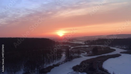 Bright sun in the clear sunset evening sky in frosty weather Bright sun on the horizon and glare on the river thaw. A drone flight over a snow-covered rural landscape to the setting sun on the horizon