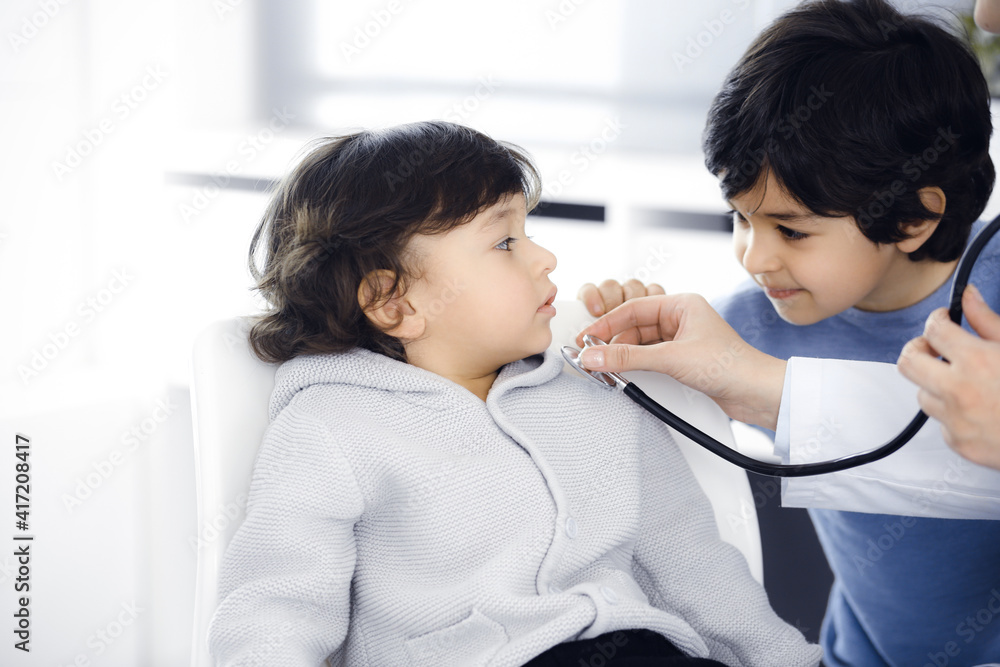 Doctor-woman examining a child patient by stethoscope. Cute arab boy and his brother at physician appointment. Medicine concept Doctor-woman examining a child patient by stethoscope. Cute arab boy and his brother at physician appointment. Medicine concept