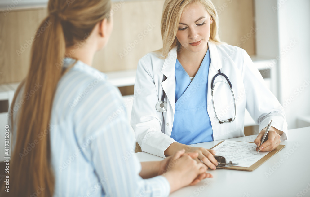 Woman-doctor and patient sitting and talking in hospital or clinic. Blonde therapist is cheerfully smiling. Medicine concept Woman-doctor and patient sitting and talking in hospital or clinic. Blonde therapist is cheerfully smiling. Medicine concept
