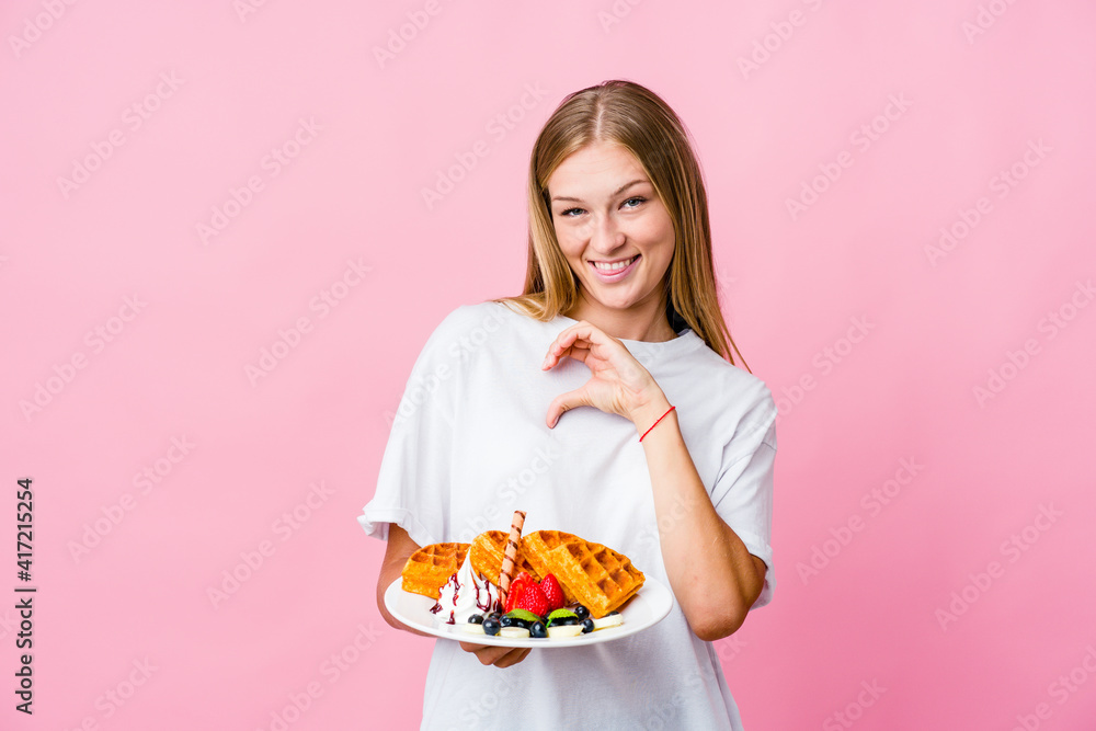 Young russian woman eating a waffle isolated smiling and showing a heart shape with hands.