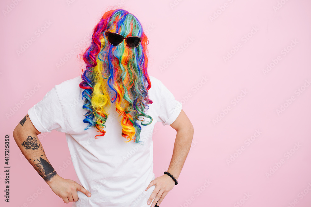 A man jokingly put on a multi-colored wig and glasses on his head, posing against a pink studio background. - obrazy, fototapety, plakaty 