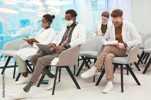 Side view at multi-ethnic group of people wearing masks and lab coats while listening to lecture on medicine in college or coworking center, copy space
