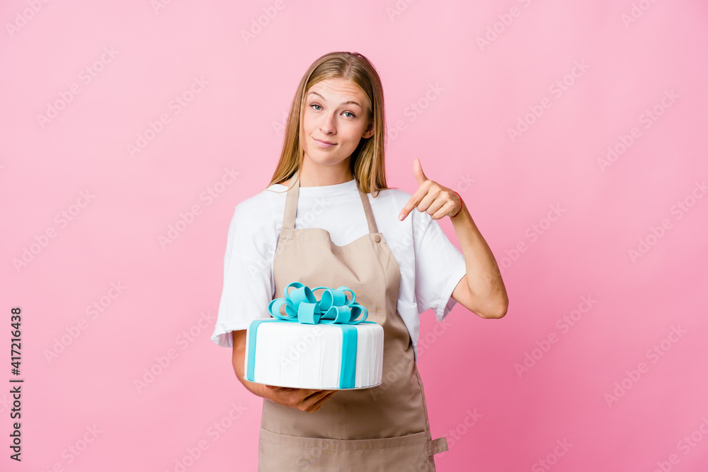 Young russian baker woman holding a delicious cake points down with fingers, positive feeling. Young russian baker woman holding a delicious cake points down with fingers, positive feeling.