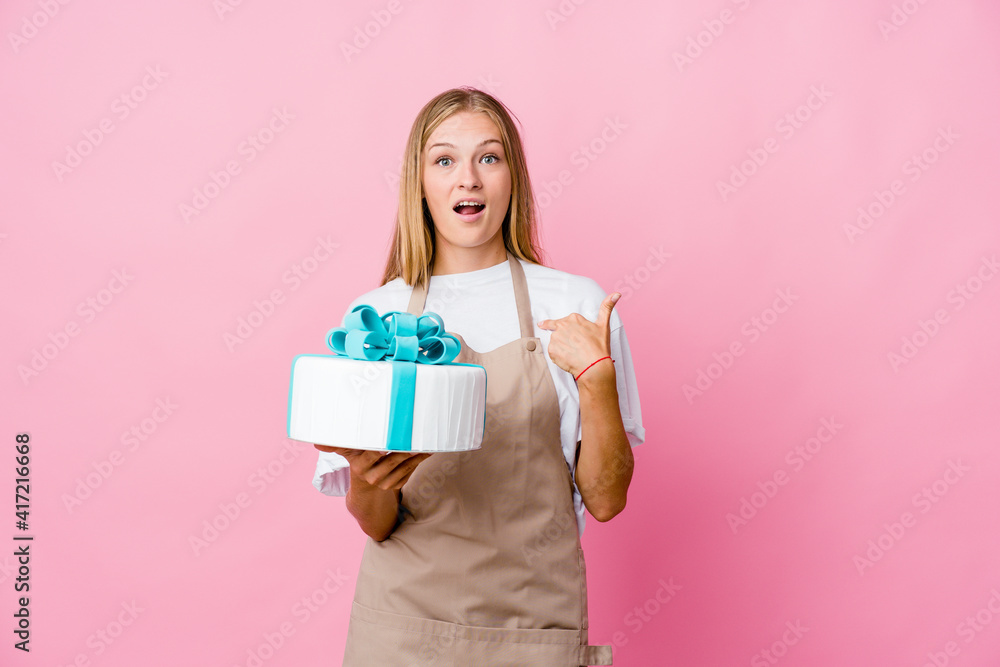 Young russian baker woman holding a delicious cake surprised pointing with finger, smiling broadly. Young russian baker woman holding a delicious cake surprised pointing with finger, smiling broadly.