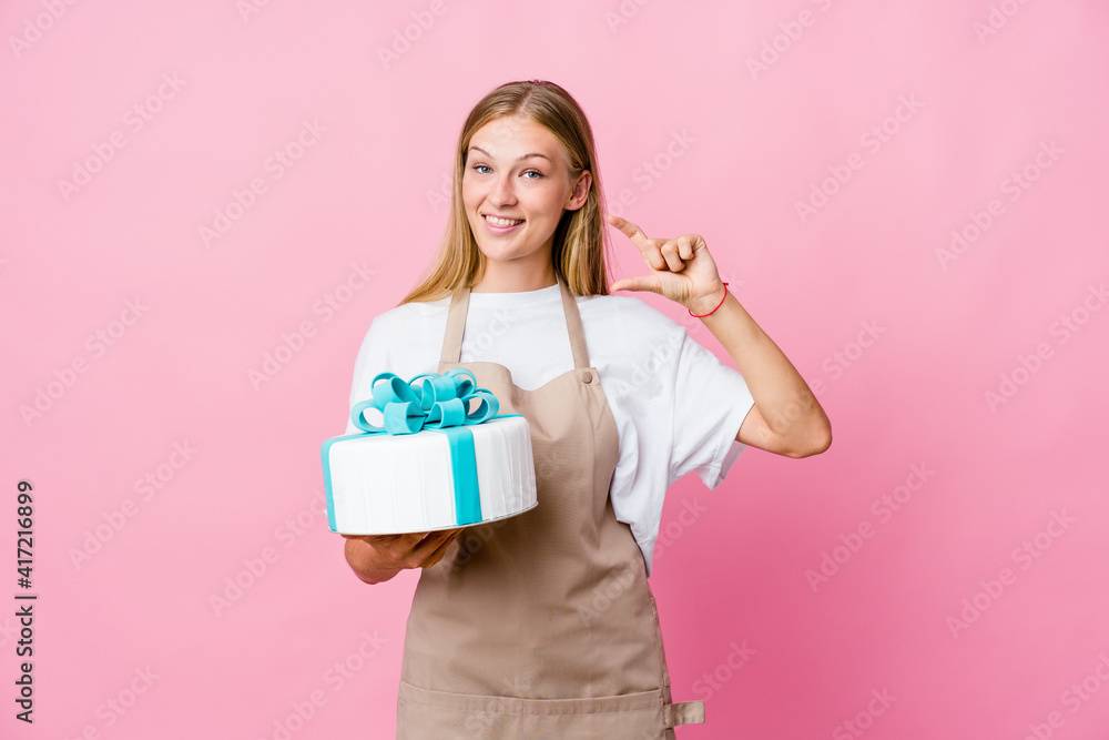 Young russian baker woman holding a delicious cake holding something little with forefingers, smiling and confident. Young russian baker woman holding a delicious cake holding something little with forefingers, smiling and confident.