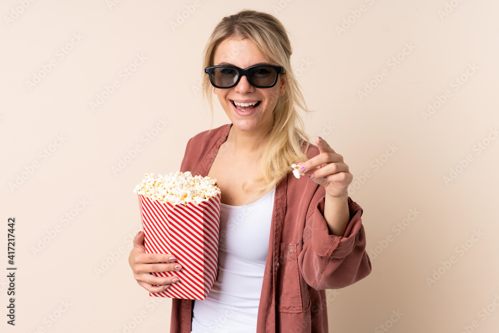 Blonde woman over isolated background with 3d glasses and holding a big bucket of popcorns while pointing front