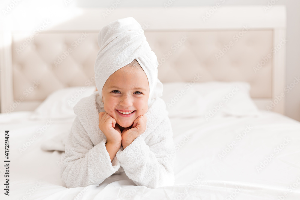 little smiling girl in a white robe with a white towel on her head is lying in bed. portrait of a child little smiling girl in a white robe with a white towel on her head is lying in bed. portrait of a child