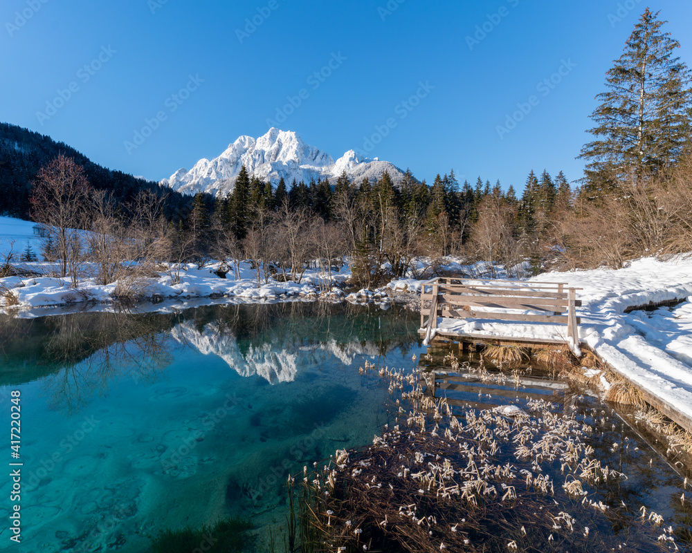 romantic path through snow along a beautiful emerald, clear volcanic lake in winter at a touristic nature reserve in the Alps. Reflection of snowcapped mountains glowing in the rising sun. no people romantic path through snow along a beautiful emerald, clear volcanic lake in winter at a touristic nature reserve in the Alps. Reflection of snowcapped mountains glowing in the rising sun. no people