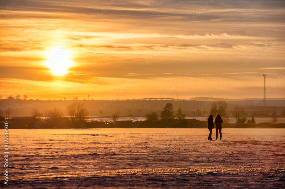 Zwei Menschen schauen sich auf einem zugefrorenen See den romantischen Sonnenuntergang an