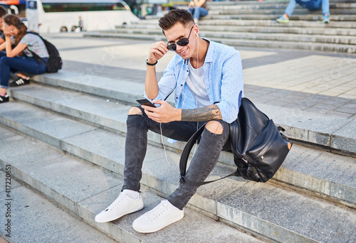 A young guy, in sunglasses and with a black backpack. He sits on the steps in the town square with telephones in his hands and looks into the frame.