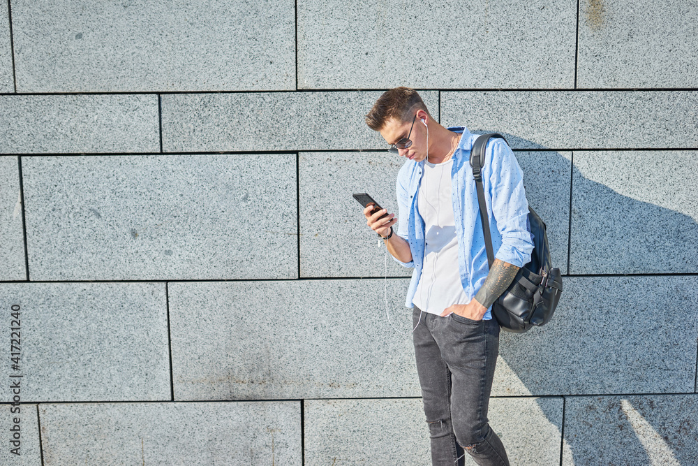 A young guy, European appearance, in sunglasses and with a black backpack. Stands near the wall and looks into the phone while.