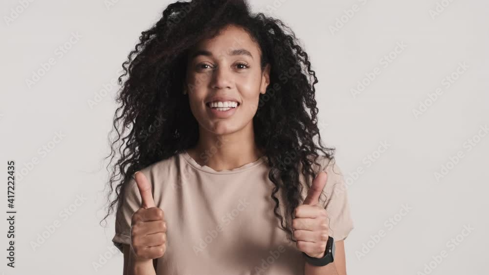 Young African American woman with dark fluffy hair keeping thumbs up and smiling on camera over white background. Like it expression Young African American woman with dark fluffy hair keeping thumbs up and smiling on camera over white background. Like it expression