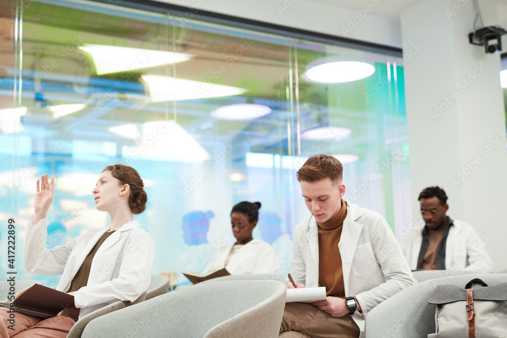 Fototapeta premium Side view at group of young people wearing lab coats while sitting in audience and listening to lecture on medicine in college or coworking center, copy space