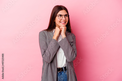 Young caucasian business woman isolated on pink background keeps hands under chin, is looking happily aside. © Asier