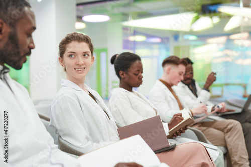 Side view at multi ethnic group of young people wearing lab coats while sitting in row in audience and listening to lecture on medicine in college or coworking center  copy space