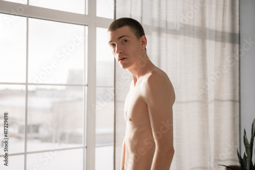 a thin guy stands by the window and looks at the camera