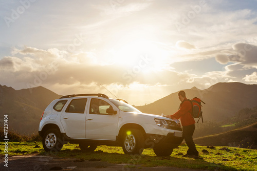 Hiker with backpack looking at a map on the hood of his off-road car at sunset. Traveler and adventurous person traces his mountain route on a map. suv vehicle.