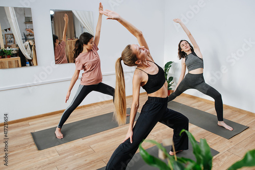 Flexible young woman practicing yoga in a group in front of an instructor. They are holding warrior asana, leaning body back with one arm sliding on the leg and the other outstretched up.