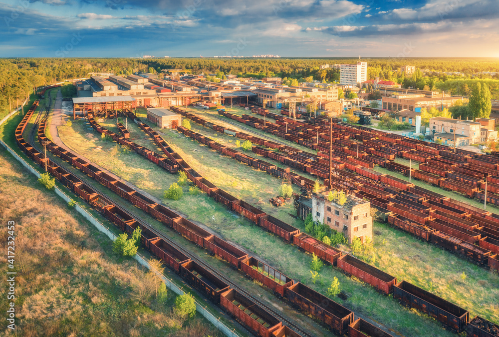 Aerial view of freight trains at sunset. Railway station. Rusty wagons on railroad. Heavy industry. Industrial landscape with train in depot, green trees, buildings, sky. Top view. Transportation 