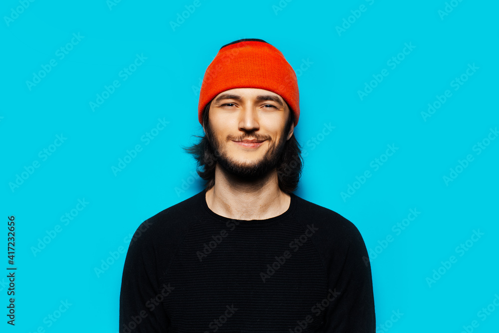 Studio portrait of young confident man, with orange beanie hat. On the background of bright blue wall. Studio portrait of young confident man, with orange beanie hat. On the background of bright blue wall.