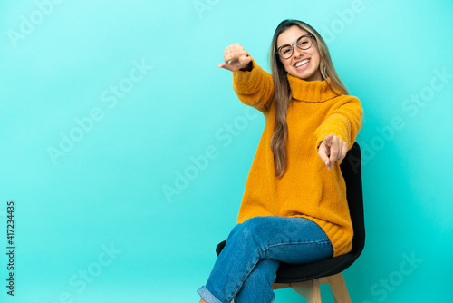 Young caucasian woman sitting on a chair isolated on blue background points finger at you while smiling © luismolinero
