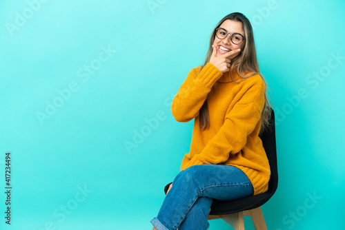 Young caucasian woman sitting on a chair isolated on blue background smiling © luismolinero