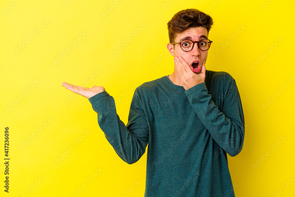 Young caucasian man isolated on yellow background holds copy space on a palm, keep hand over cheek. Amazed and delighted. Young caucasian man isolated on yellow background holds copy space on a palm, keep hand over cheek. Amazed and delighted.