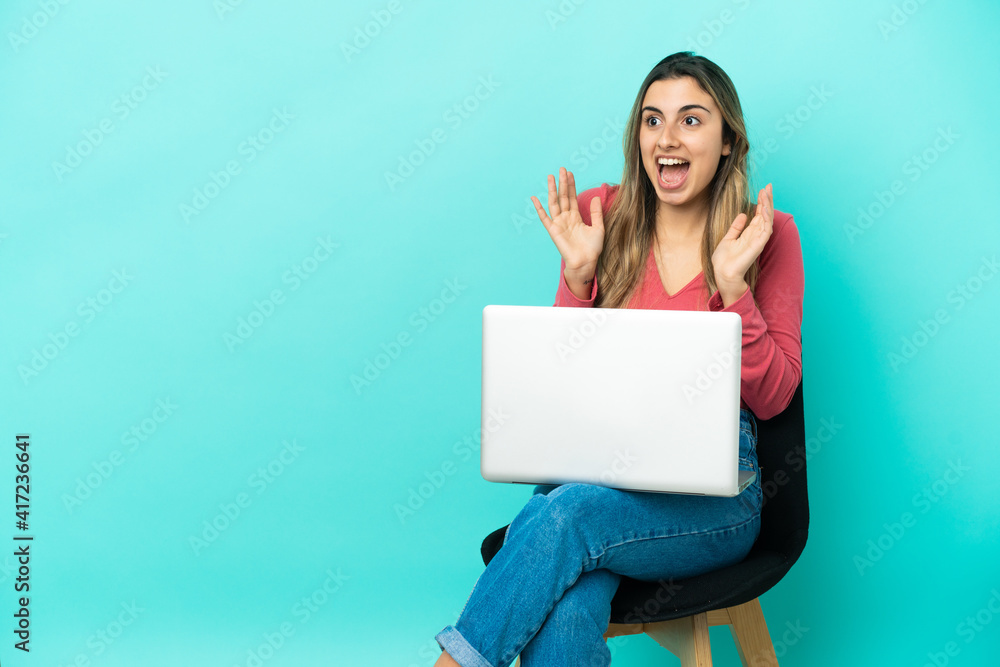 Young caucasian woman sitting on a chair with her pc isolated on blue background with surprise facial expression Young caucasian woman sitting on a chair with her pc isolated on blue background with surprise facial expression