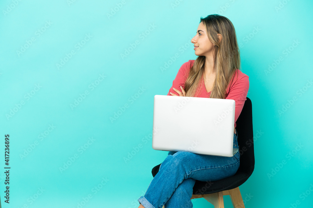 Young caucasian woman sitting on a chair with her pc isolated on blue background in lateral position Young caucasian woman sitting on a chair with her pc isolated on blue background in lateral position