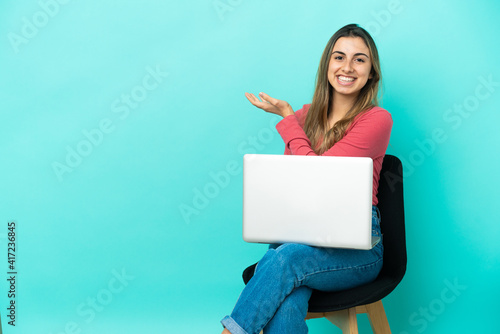 Young caucasian woman sitting on a chair with her pc isolated on blue background presenting an idea while looking smiling towards © luismolinero