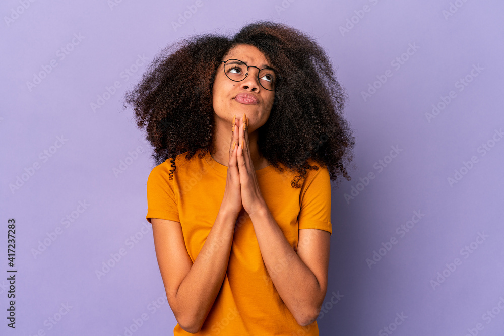 Young african american curly woman isolated on purple background holding hands in pray near mouth, feels confident. Young african american curly woman isolated on purple background holding hands in pray near mouth, feels confident.