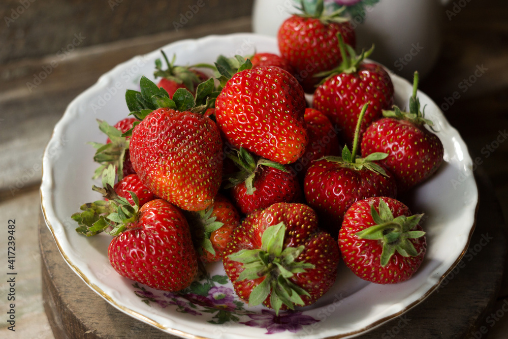 Ripe strawberries in a white old plate, near a rose. Brown wooden background Ripe strawberries in a white old plate, near a rose. Brown wooden background