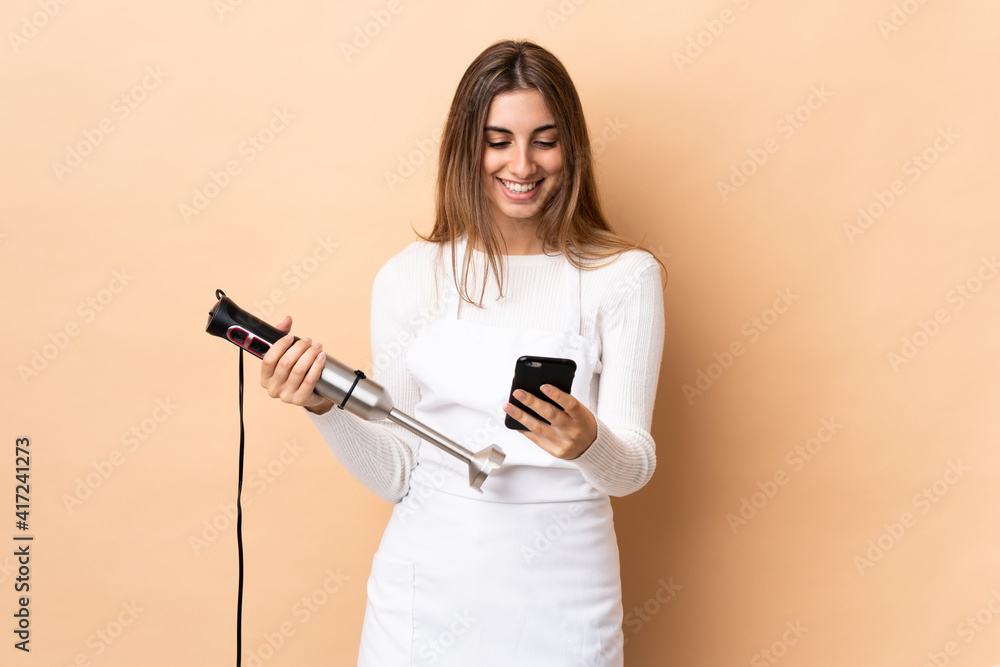 Woman using hand blender over isolated background sending a message with the mobile Woman using hand blender over isolated background sending a message with the mobile