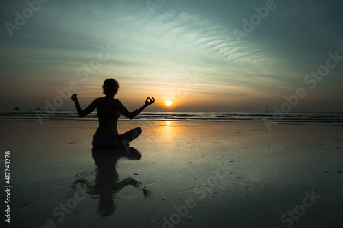 An woman sitting on the beach during sunset and meditating in yoga pose.