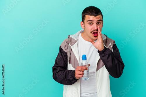 Young sporty caucasian man drinking water isolated on blue background is saying a secret hot braking news and looking aside