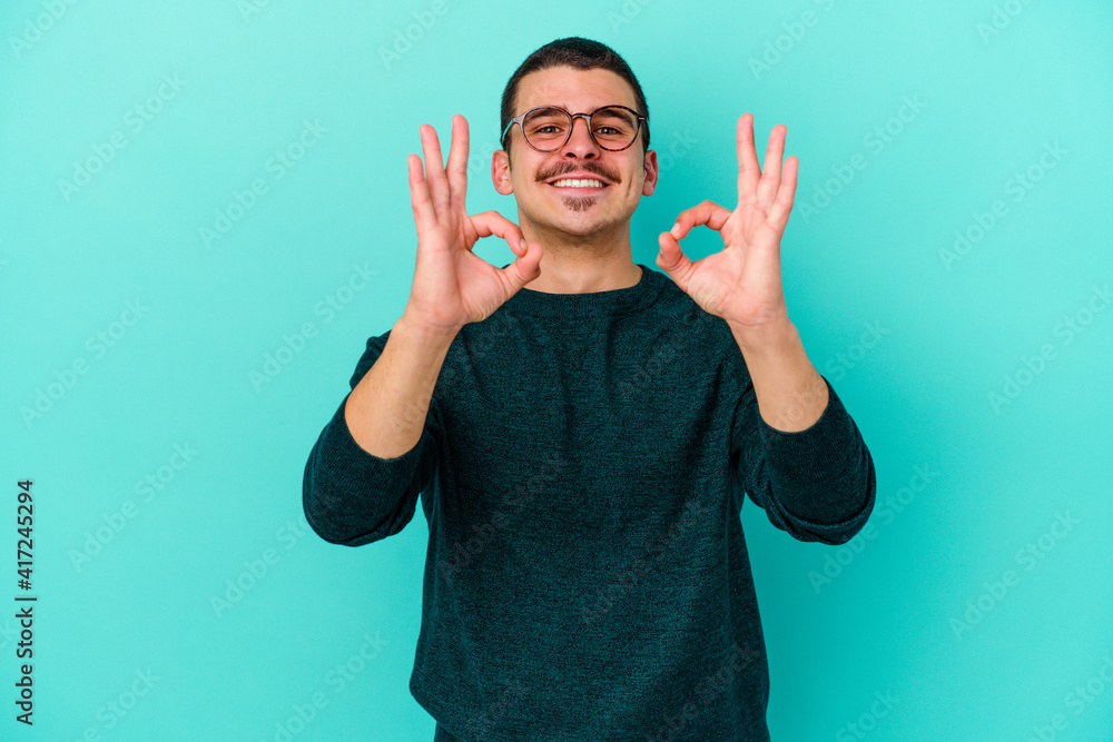 Young caucasian man isolated on blue background cheerful and confident showing ok gesture. Young caucasian man isolated on blue background cheerful and confident showing ok gesture.