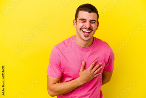 Young caucasian cool man isolated on yellow background laughs out loudly keeping hand on chest.