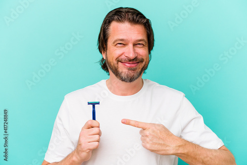 Middle age dutch man holding a razor blade isolated on blue background smiling and pointing aside, showing something at blank space.