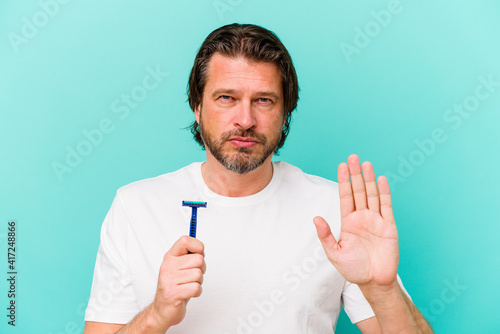 Middle age dutch man holding a razor blade isolated on blue background standing with outstretched hand showing stop sign, preventing you.